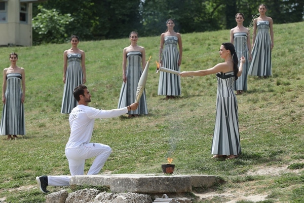 扮演最高女祭司的演員瑪麗·米娜（前右）點(diǎn)燃第一棒火炬手——希臘運(yùn)動員、東京奧運(yùn)會男子賽艇項目金牌得主斯特凡諾斯·恩圖斯科斯手中的火炬。新華社記者 趙丁喆 攝