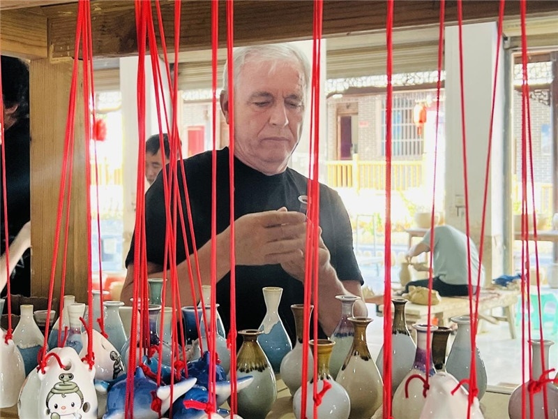 鄉(xiāng)愁如同一根根紅線，將游子與故鄉(xiāng)永遠地牽連到一起?；逄账囄幕瘎?chuàng)意一條街的母氏陶藝館內，紅線吊著陶瓶，思緒游走萬里。摩爾多瓦“信息標簽”通訊社社長亞歷山大·塔納斯端詳著館內展示的陶藝作品，并挑選出心儀的小擺件帶回摩爾多瓦，也帶走花茂村的“鄉(xiāng)愁”。（張亞欣攝）